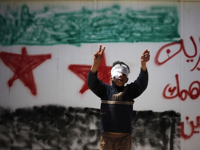 Tại trại tị nạn ở Jordan, một cậu bé Syria bị thương ở mắt đứng trước bức tường có dòng chữ tiếng Ảrập: “Syria, đừng lo, chúng tôi sẽ quay về”.